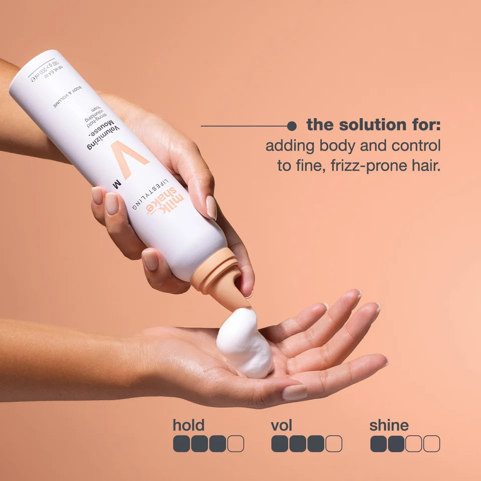 A hand holds a white bottle of MIlk_shake Hair Volumizing Hair Enhancer, squeezing white foam into another hand, against a peach background with product text and hold/vol/shine ratings.